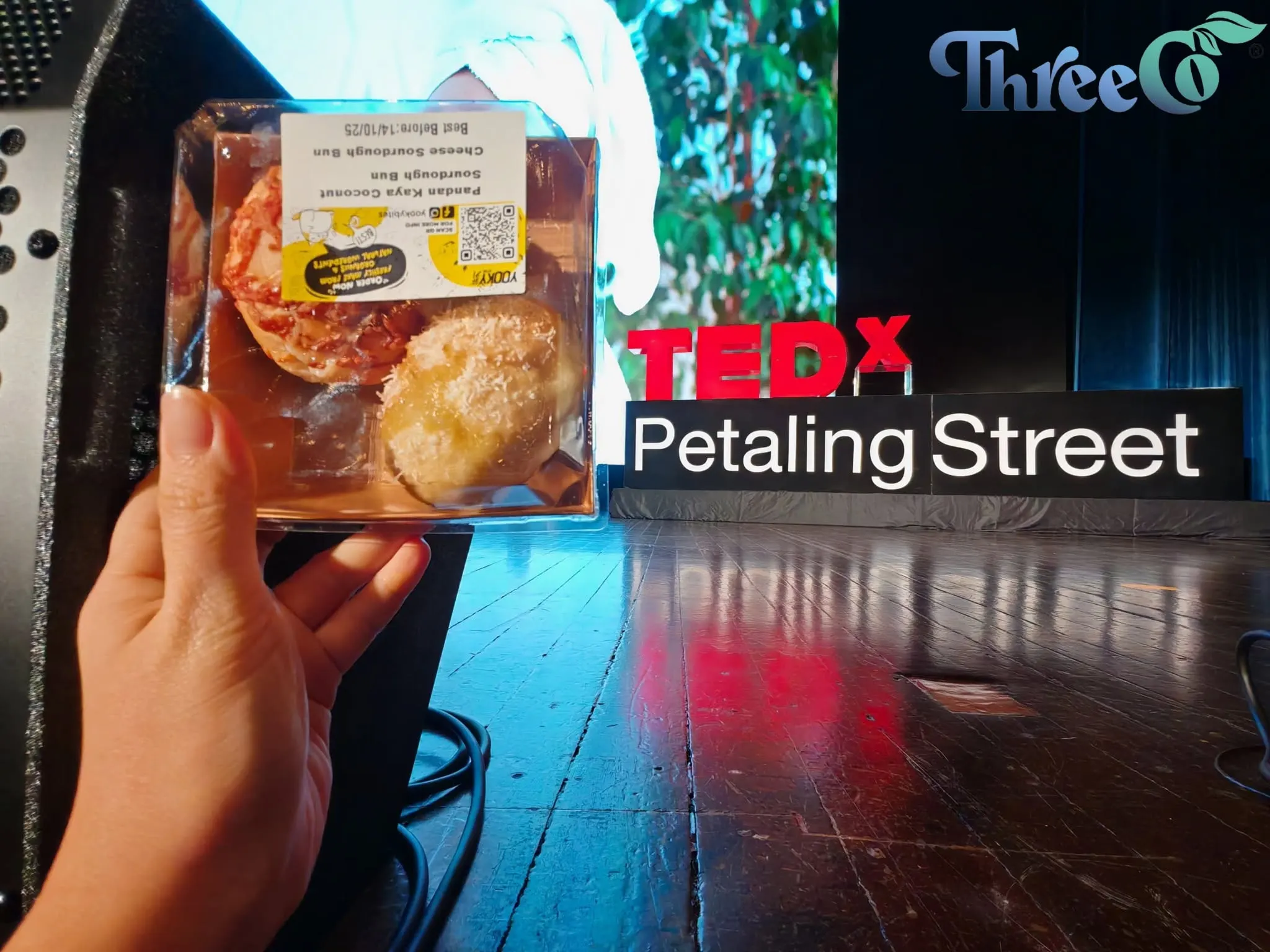 A box of sourdough buns held up in front of the TEDxPetalingStreet stage sign at the 2025 annual conference